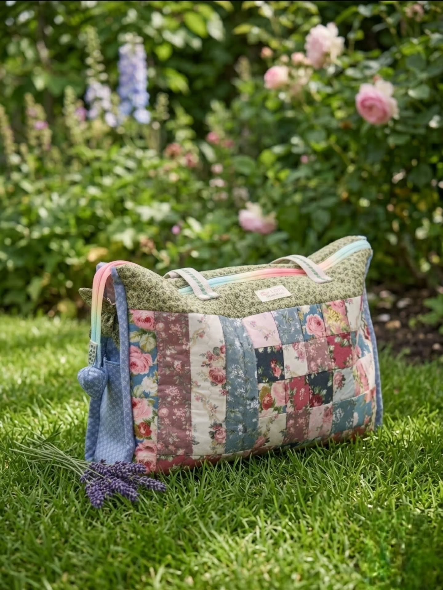 Bolso de viaje de patchwork acolchado con telas florales rosas y azules y asas multicolor, sobre el césped de un jardín